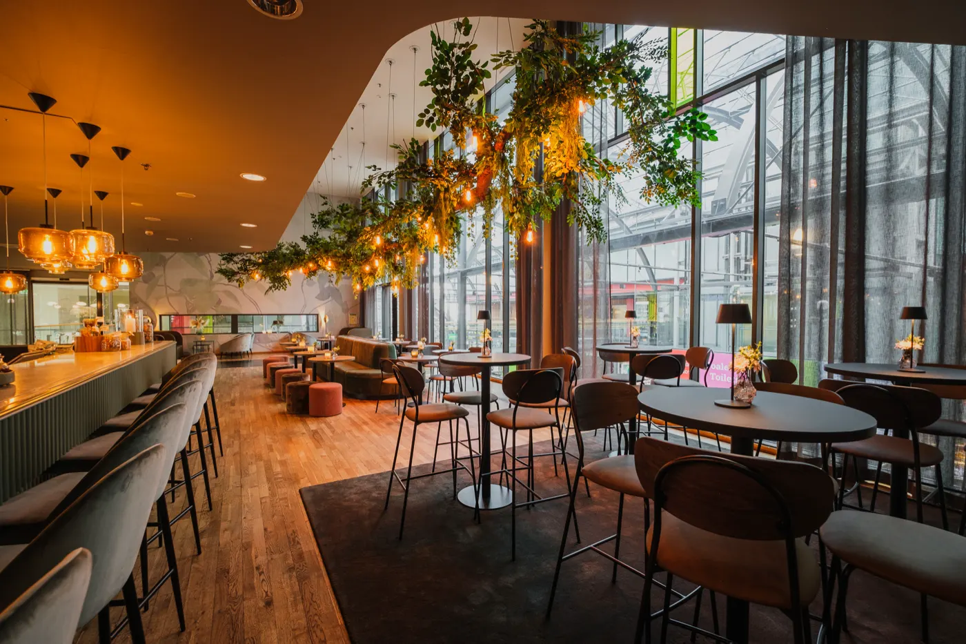 Ljus lokal med lång bardisk, hängande grönska och höga fönster mot gatan