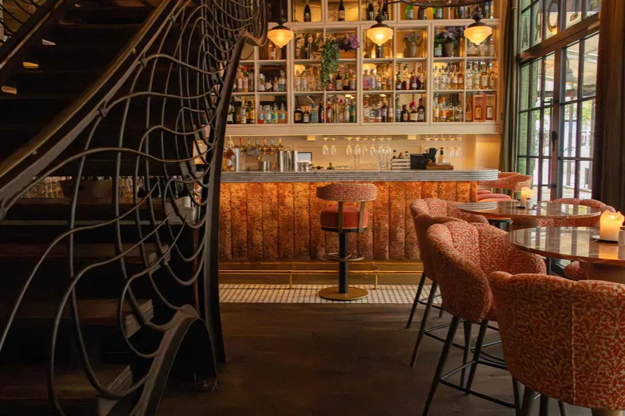 Bar med flaskhyllor och rostfärgade barstolar bredvid en svängd jugendtrappa