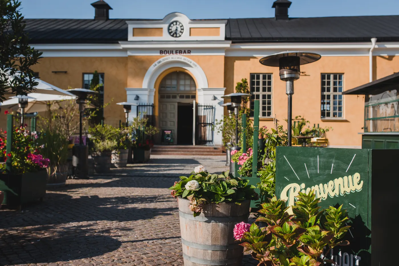 Gul ridhusbyggnad med klocka över porten, kullersten, blomkrukor och parasoller